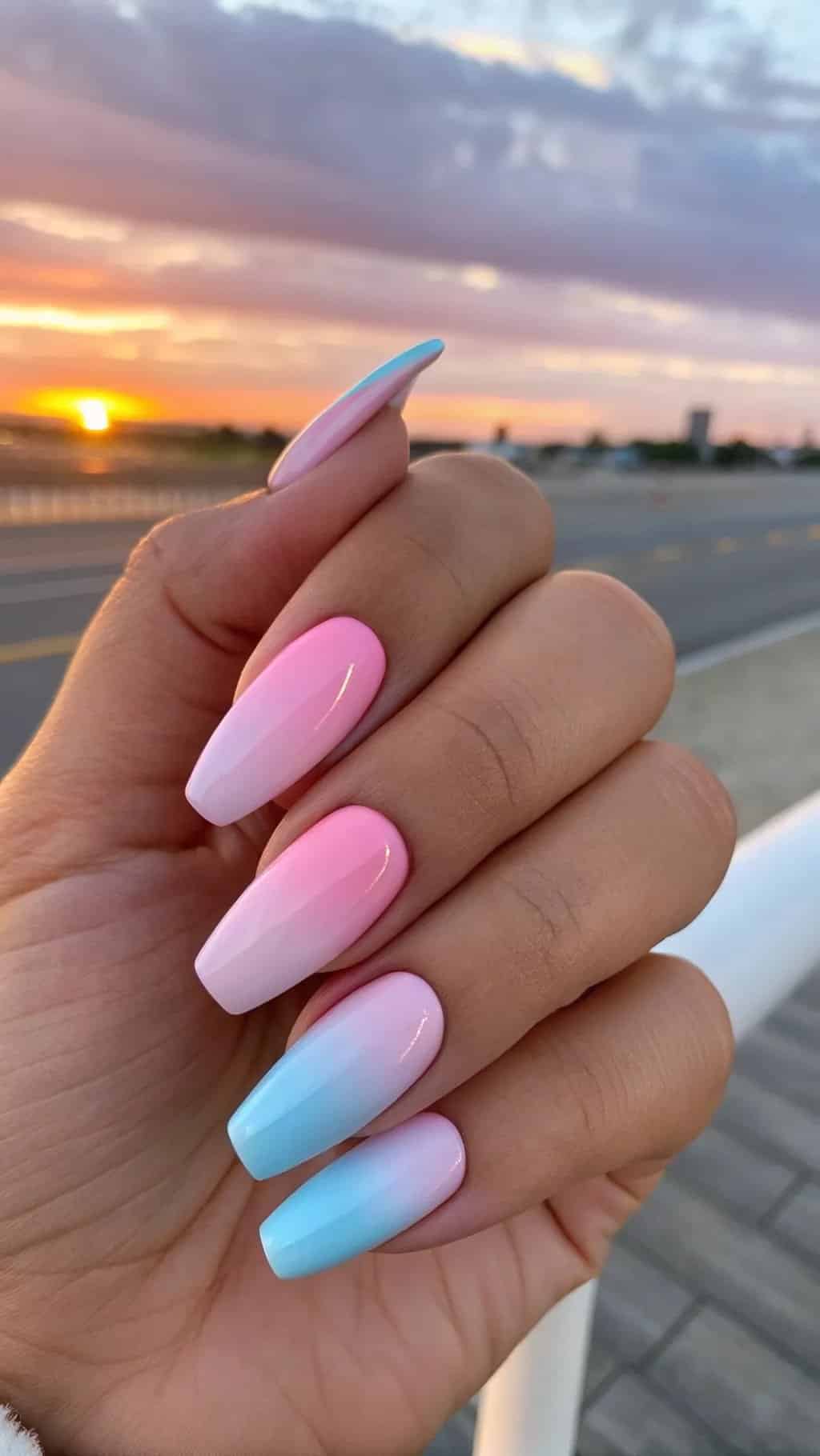 9 Trendy Summer Nail Designs for 2026 5 A hand displaying pastel ombre nails, blending pink and blue shades with a beautiful sunset in the background.