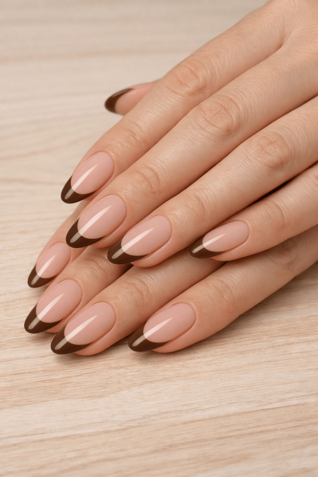 Nude almond nails with brown French tips on a wooden surface.